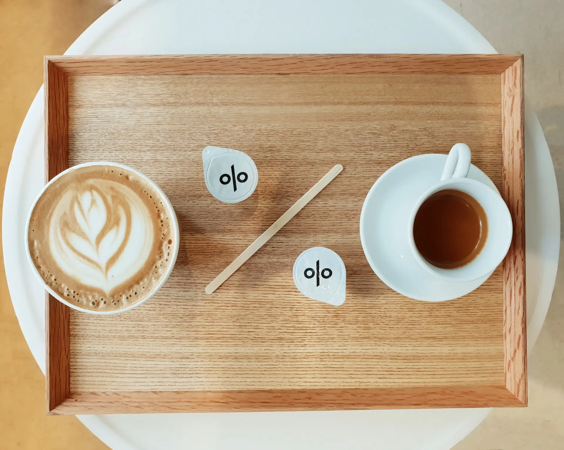 Drewniana taca z ceramiczną filiżanką espresso i latte, dwiema paczkami cukru i drewnianymi mieszadełkami. Całość układa się w kształt znaku procenta.